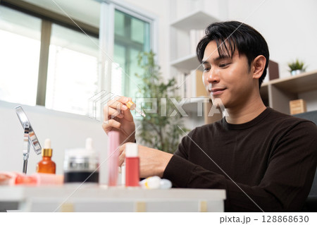 Skincare Routine. Young man smiling while applying serum in bright room. Skincare Routine. Young man smiling while applying serum in bright room. 128868630