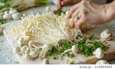 Close Up female hands preparing fresh enoki mushrooms with chopped parsley on a wooden cutting board. Cooking process, vegetarian ingredients and kitchen vibes Close Up female hands preparing fresh enoki mushrooms with chopped parsley on a wooden cutting board. Cooking process, vegetarian ingredients and kitchen vibes 128868904