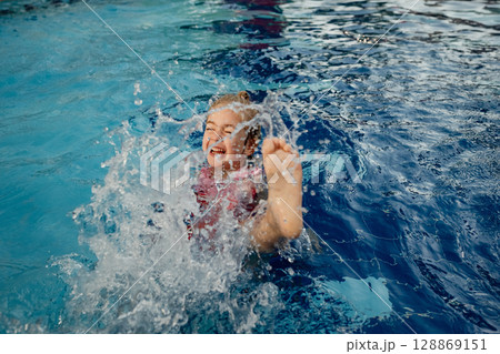 Child enjoys playful splashing while swimming in a bright blue pool on a sunny day 128869151