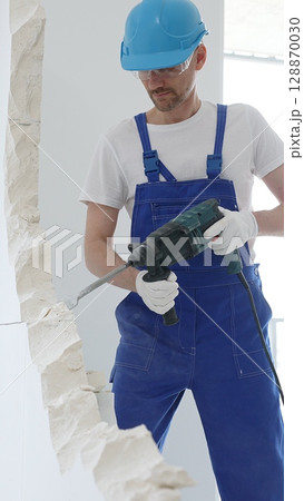 Man construction worker wearing blue overalls, hard hat and protective white gloves, is demolishing white wall with rotary hammer drill, generating dust, close up horizontal view. Renovation concept 128870030