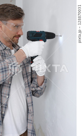 Middle aged man worker wearing grey checked shirt and white gloves, is drilling hole in a wall with cordless drill during home renovation work. Portrait view Middle aged man worker wearing grey checked shirt and white gloves, is drilling hole in a wall with cordless drill during home renovation work. Portrait view 128870031