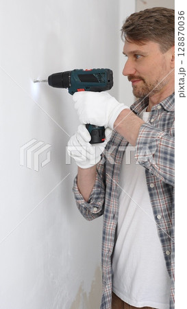 Middle aged male professional worker wearing grey checked shirt and white gloves, is drilling hole in a wall with cordless drill during home renovation work. Vertical portrait view 128870036