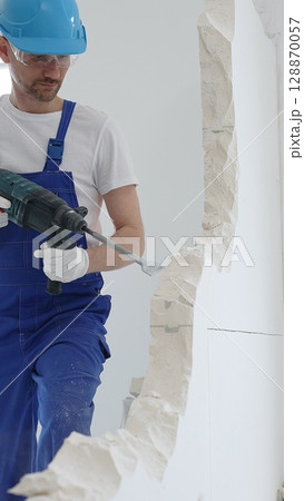 Male construction worker wearing blue overalls, protective helmet and white gloves, is demolishing white wall with rotary hammer drill, generating dust, close up horizontal view Male construction worker wearing blue overalls, protective helmet and white gloves, is demolishing white wall with rotary hammer drill, generating dust, close up horizontal view 128870057
