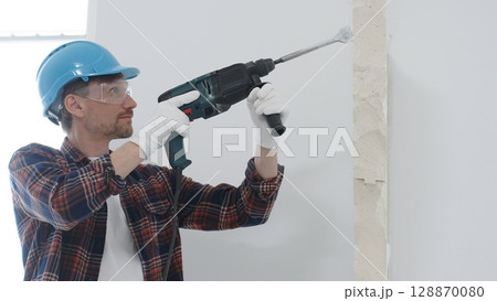 Male construction worker wearing red checkered shirt, blue protective helmet and gloves, is demolishing white wall with rotary hammer drill, generating dust, portrait view. Renovation concept Male construction worker wearing red checkered shirt, blue protective helmet and gloves, is demolishing white wall with rotary hammer drill, generating dust, portrait view. Renovation concept 128870080