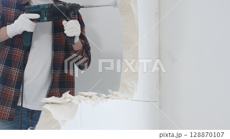 Close up of unknown male construction worker wearing red checkered shirt jeans and protective gloves, is demolishing white wall with rotary hammer drill, generating dust. Renovation concept Close up of unknown male construction worker wearing red checkered shirt jeans and protective gloves, is demolishing white wall with rotary hammer drill, generating dust. Renovation concept 128870107