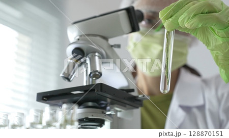 Female scientist in green protective mask and gloves holding test tube with plant sample, researching botany and medical applications using a microscope. Medicine, health care and science concept 128870151