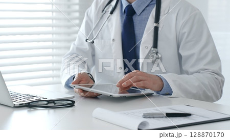 Male doctor in lab coat and stethoscope using digital tablet while sitting at table in medical office with laptop, glasses and medical chart nearby. Medicine and healthcare concept 128870170