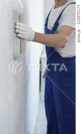 Man construction worker wearing protective gloves and blue construction coveralls, spreading plaster smoothly across wall using professional drywall taping knife Man construction worker wearing protective gloves and blue construction coveralls, spreading plaster smoothly across wall using professional drywall taping knife 128870171