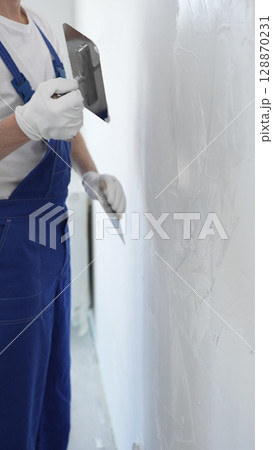 Professional construction worker wearing blue work overall is using a finishing trowel to carefully apply plaster on a wall, demonstrating expertise in home renovation Professional construction worker wearing blue work overall is using a finishing trowel to carefully apply plaster on a wall, demonstrating expertise in home renovation 128870231