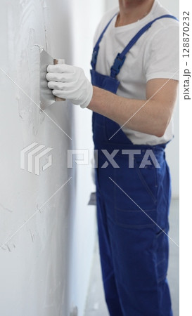 Unknown man construction worker wearing gloves and blue overalls is using a putty knife to apply plaster on a white wall, performing home renovation and improvement 128870232