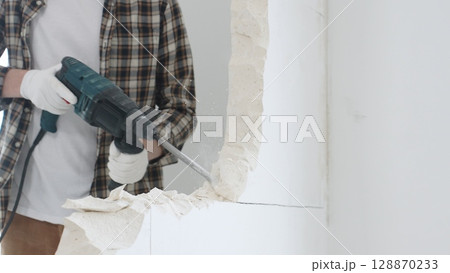 Close up of unrecognizable male construction worker wearing beige checkered shirt and protective gloves, is demolishing white wall with rotary hammer drill, generating dust, close up view. Renovation Close up of unrecognizable male construction worker wearing beige checkered shirt and protective gloves, is demolishing white wall with rotary hammer drill, generating dust, close up view. Renovation 128870233