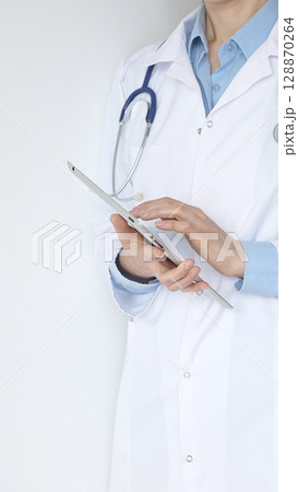 Unknown female doctor, wearing white coat over a blue blouse and a stethoscope, is using a digital tablet, suggesting innovative healthcare practices in medicine. Health care and medicine concept 128870264