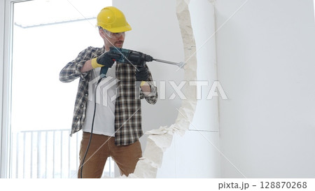 Male construction worker wearing beige checkered shirt, yellow hard hat and protective gloves, is demolishing white wall with rotary hammer drill, generating dust, medium long shot. Renovation 128870268