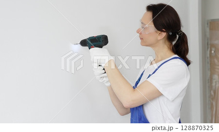 Woman worker wearing blue overall, safety glasses and gloves, is using cordless drill to make hole in white wall during renovation work. Horizontal portrait view 128870325