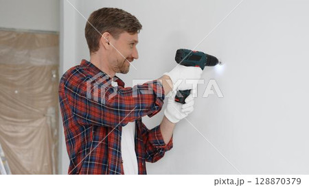 Middle aged male worker wearing red checked shirt and white gloves, is drilling hole in a wall with cordless drill during home renovation work. Portrait view Middle aged male worker wearing red checked shirt and white gloves, is drilling hole in a wall with cordless drill during home renovation work. Portrait view 128870379