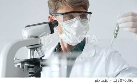 Adult male scientist in mask and gloves holding test tube with green plant sample, conducting laboratory research with a microscope. Medicine, healthcare and science concept Adult male scientist in mask and gloves holding test tube with green plant sample, conducting laboratory research with a microscope. Medicine, healthcare and science concept 128870380