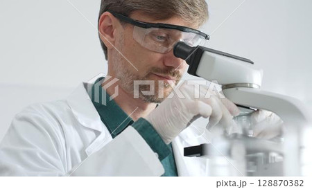 Senior man scientist wearing protective glasses and lab coat analyzing samples with a microscope in laboratory setting. Medicine, health care and science concept Senior man scientist wearing protective glasses and lab coat analyzing samples with a microscope in laboratory setting. Medicine, health care and science concept 128870382