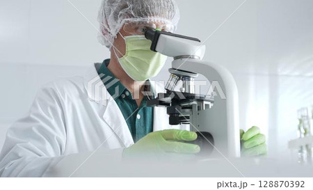 Medical scientist wearing protective mask, cap and green gloves using microscope doing scientific research in laboratory, developing vaccine or medication. Medicine, healthcare and science concept Medical scientist wearing protective mask, cap and green gloves using microscope doing scientific research in laboratory, developing vaccine or medication. Medicine, healthcare and science concept 128870392