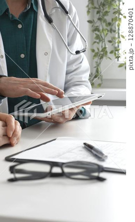 Doctor man in lab coat over a green shirt, and stethoscope is using tablet while consulting patient in modern medical office. Medicine and health care concept 128870401