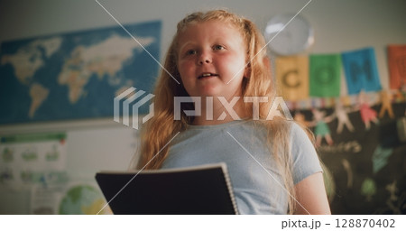 Primary School Girl with Notebook Speaking, Showcasing Knowledge of Geography in Front of Classroom 128870402