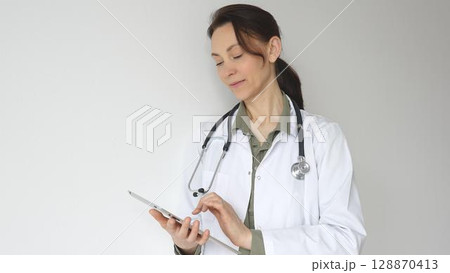 Doctor woman wearing white medical coat over green blouse, stethoscope, is reviewing patient records on digital tablet, standing in medical workspace with natural lighting. Medicine concept 128870413