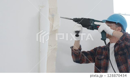 Man construction worker wearing red checkered shirt, blue hard hat and protective gloves, is demolishing white wall with rotary hammer drill, generating dust, portrait view. Renovation concept 128870434
