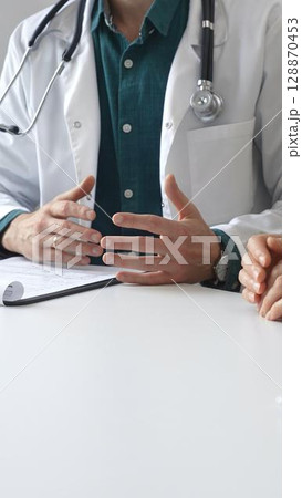 Doctor man wearing medical coat over green shirt, and stethoscope is gesturing with hands while explaining diagnosis to patient during medical consultation in clinic office. Medicine and health care Doctor man wearing medical coat over green shirt, and stethoscope is gesturing with hands while explaining diagnosis to patient during medical consultation in clinic office. Medicine and health care 128870453