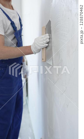 Man construction worker wearing protective gloves and blue construction coveralls, spreading plaster smoothly across wall using professional drywall taping knife 128870462