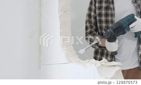 Close up of unrecognizable male construction worker wearing beige checkered shirt and protective gloves, is demolishing white wall with rotary hammer drill, generating dust, close up view. Renovation Close up of unrecognizable male construction worker wearing beige checkered shirt and protective gloves, is demolishing white wall with rotary hammer drill, generating dust, close up view. Renovation 128870573