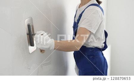Professional female construction worker is using a finishing trowel to carefully apply plaster on a wall, demonstrating expertise in home renovation and interior finishing 128870653