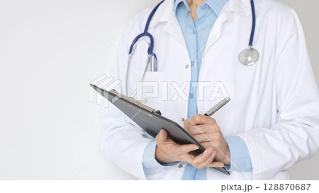 Close up of doctor woman wearing stethoscope and white coat over blue blouse is standing and writing on a black clipboard, representing medical expertise and patient care. Medicine concept Close up of doctor woman wearing stethoscope and white coat over blue blouse is standing and writing on a black clipboard, representing medical expertise and patient care. Medicine concept 128870687