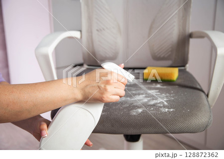 A woman's hand with a cleaning agent sprays a spray on the dirty upholstery of a computer chair. Concept of dry cleaning at home, industry 128872362