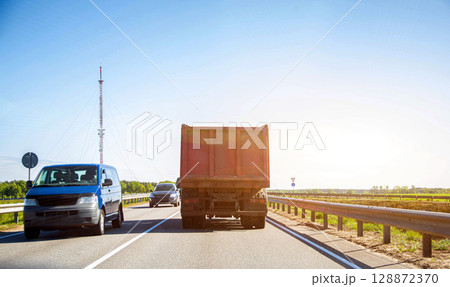 Dump truck on a country road against the background of cars, sun and sky. Transportation of bulk cargo. Copy space for text Dump truck on a country road against the background of cars, sun and sky. Transportation of bulk cargo. Copy space for text 128872370