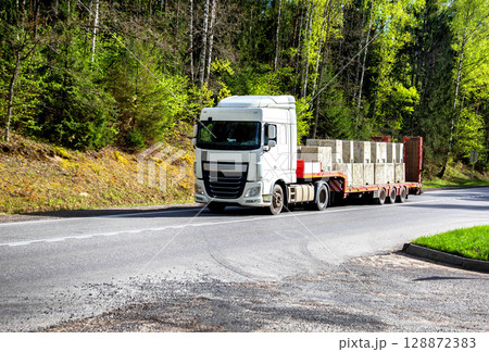 Modern truck with a lowboy semitrailer transports concrete blocks along a country road against the backdrop of a forest in summer. Beautiful nature, industry. Copy space for text Modern truck with a lowboy semitrailer transports concrete blocks along a country road against the backdrop of a forest in summer. Beautiful nature, industry. Copy space for text 128872383