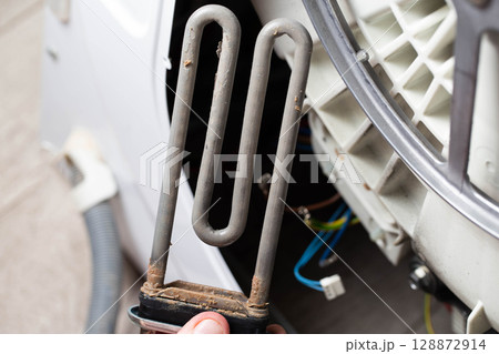 An electric heating element with scale and deposits against the background of a washing machine. Concept of repair and replacement of spare parts, close-up 128872914