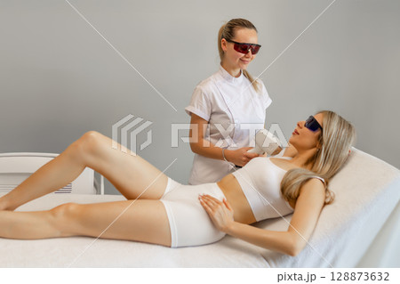 Hair removal procedure. Cropped shot of young fit woman client receiving laser hair removal procedure on body in beauty clinic. Women beauty and cosmetology concept Hair removal procedure. Cropped shot of young fit woman client receiving laser hair removal procedure on body in beauty clinic. Women beauty and cosmetology concept 128873632