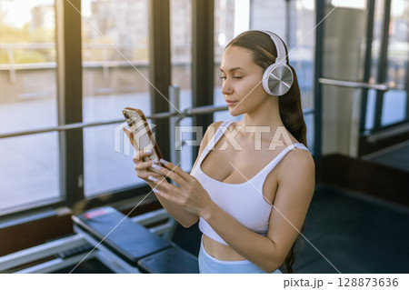 Portrait of young sportswoman with smartphone listening to music in gym. 128873636