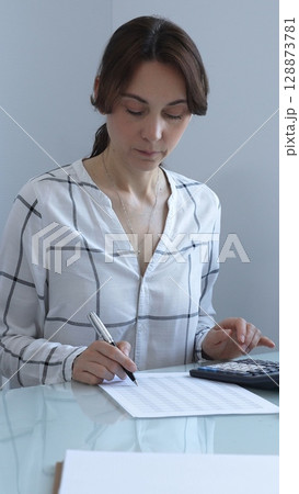 A professional woman calculates finances, checks documents, and accurately and carefully tracks business expenses at a glass table. Audit and taxes in business. 128873781