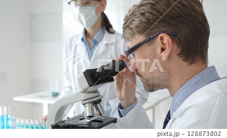 A male researcher examines a sample under a microscope, a female lab technician in a protective mask stands nearby, both in white lab coats during the study. Medicine and Science A male researcher examines a sample under a microscope, a female lab technician in a protective mask stands nearby, both in white lab coats during the study. Medicine and Science 128873802