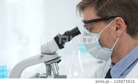 Scientist wearing mask and gloves using microscope doing scientific research in a laboratory with test tubes containing blue liquid. Medicine, healthcare and science concept 128873818