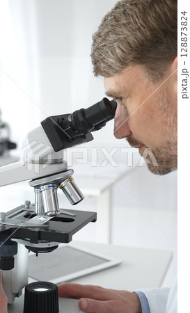 A man scientist carefully observing through a microscope in a brightly lit laboratory, conducting research for developing new medicines and treatments. Medicine and science concept 128873824