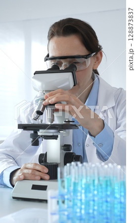 Female scientist examining microscope slide, wearing protective gear in laboratory, researching pharmaceutical breakthrough with blue tinted test tube specimens Female scientist examining microscope slide, wearing protective gear in laboratory, researching pharmaceutical breakthrough with blue tinted test tube specimens 128873837