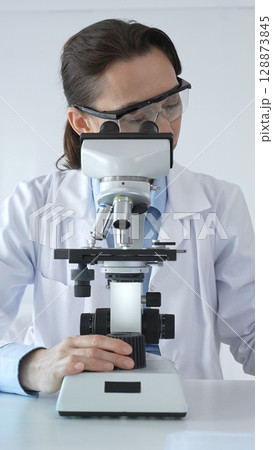 Female scientist wearing lab coat and protective glasses using microscope, analyzing samples in test tubes in modern laboratory, conducting scientific research 128873845