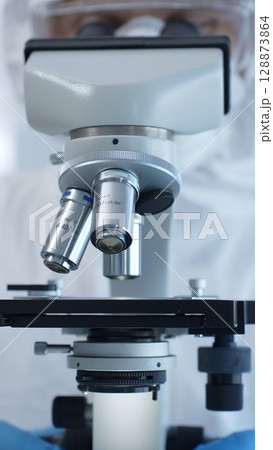 Close up of medical virologist in a hazmat suit, mask, and gloves, diligently working at a microscope in a laboratory researching viruses. Medicine and science concept Close up of medical virologist in a hazmat suit, mask, and gloves, diligently working at a microscope in a laboratory researching viruses. Medicine and science concept 128873864