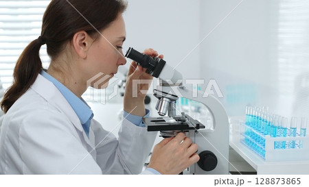 Female scientist examining microscope slide, analyzing chemical sample in sterile laboratory environment, performing precision medical research. Medicine and science concept Female scientist examining microscope slide, analyzing chemical sample in sterile laboratory environment, performing precision medical research. Medicine and science concept 128873865