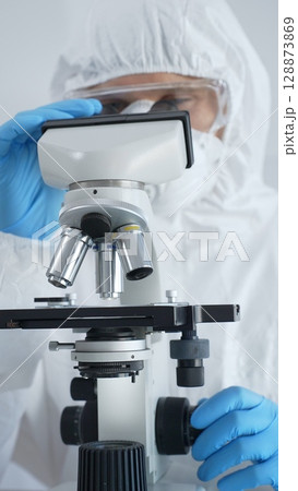 Male virologist in a hazmat suit, mask, and blue gloves, diligently working at a microscope in a laboratory researching viruses. Medicine, science concept 128873869