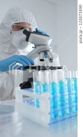 Scientific researcher wearing protective gear adjusting microscope, analyzing lab samples with precision opposite lab tubes in laboratory environment. Medicine, healthcare and science concept 128873890