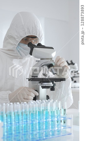 A medical virologist in protective gear examines viral samples under a microscope while conducting research in a laboratory setting A medical virologist in protective gear examines viral samples under a microscope while conducting research in a laboratory setting 128873893