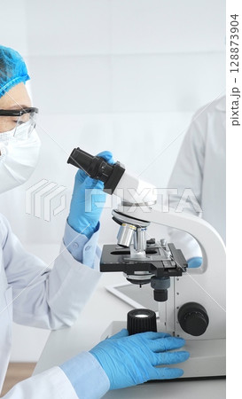 Two scientists in lab coats, face masks, and protective glasses conducting research with a microscope in a laboratory. Medicine and science concept Two scientists in lab coats, face masks, and protective glasses conducting research with a microscope in a laboratory. Medicine and science concept 128873904