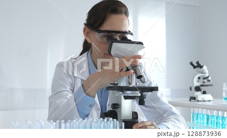 Female scientist wearing lab coat and protective glasses using microscope, analyzing samples in test tubes in modern laboratory, conducting scientific research 128873936